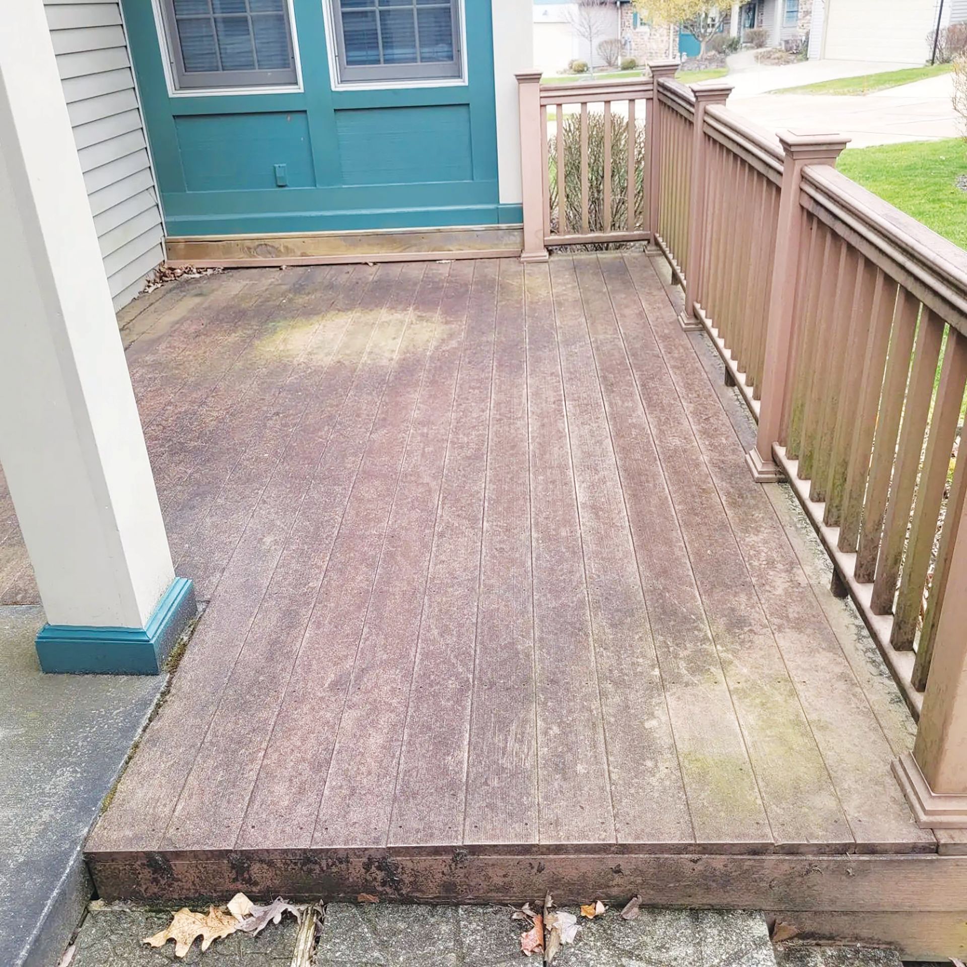 A weathered wooden deck with a teal house exterior, railing, and stairs, viewed from a slightly elevated angle.