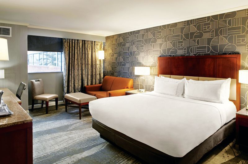 A modern hotel room featuring a king-size bed with white bedding, an orange armchair, a small ottoman, and a desk.