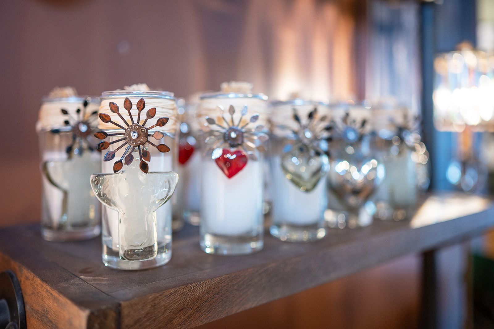 A row of small, clear glass candle holders, decorated with metal starburst accents and small hearts, on a wooden shelf.
