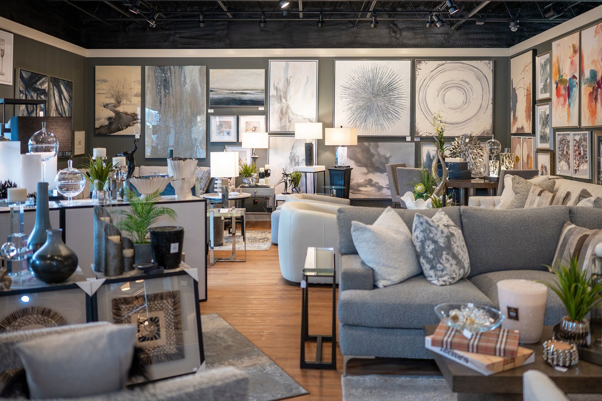 A furniture showroom featuring gray sofas, end tables, lamps, and various framed wall art.