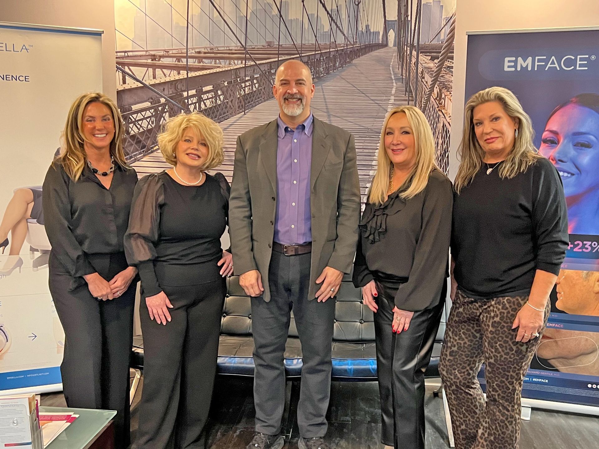 Five people stand side-by-side in front of a bridge-themed backdrop and EmFace branding in an indoor office setting.