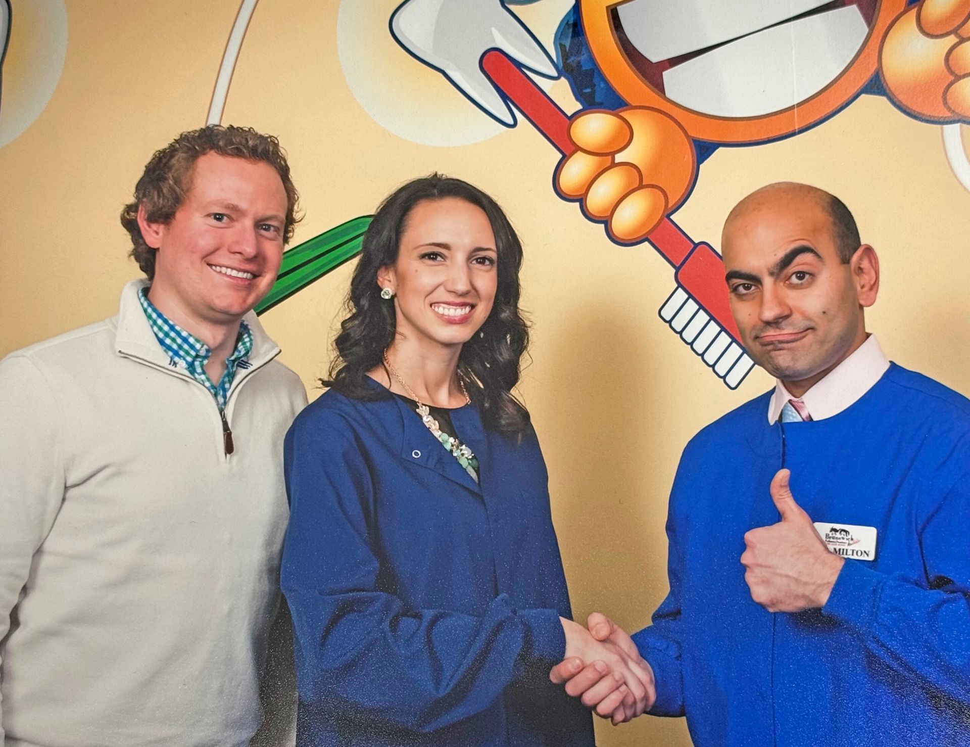 Two people shake hands while a third stands beside them in front of a colorful wall graphic featuring a toothbrush.