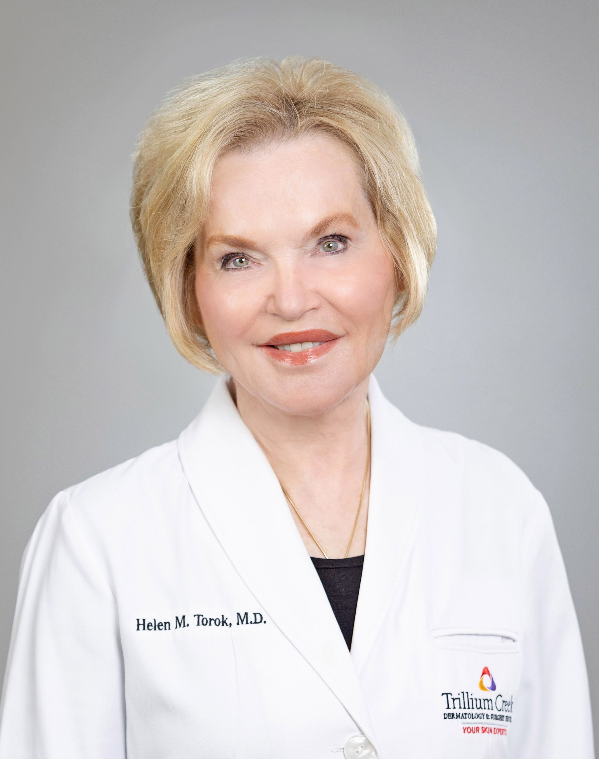 Headshot of a person in a white medical coat with a professional logo, smiling against a light gray background.