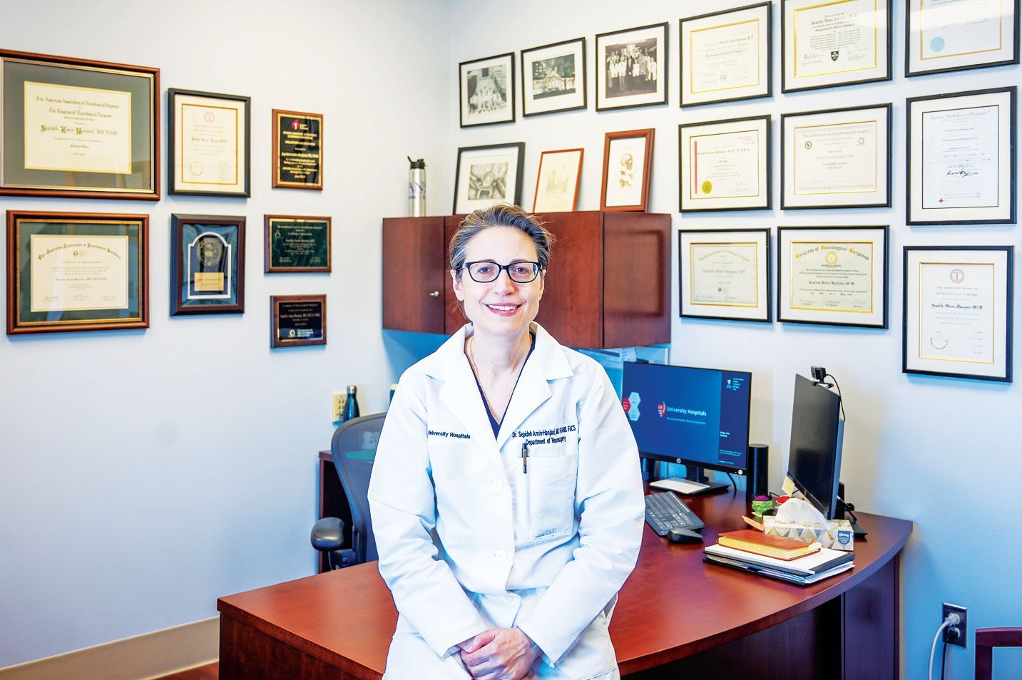 A professional sitting at a desk in a medical office, surrounded by numerous framed certificates on the wall.