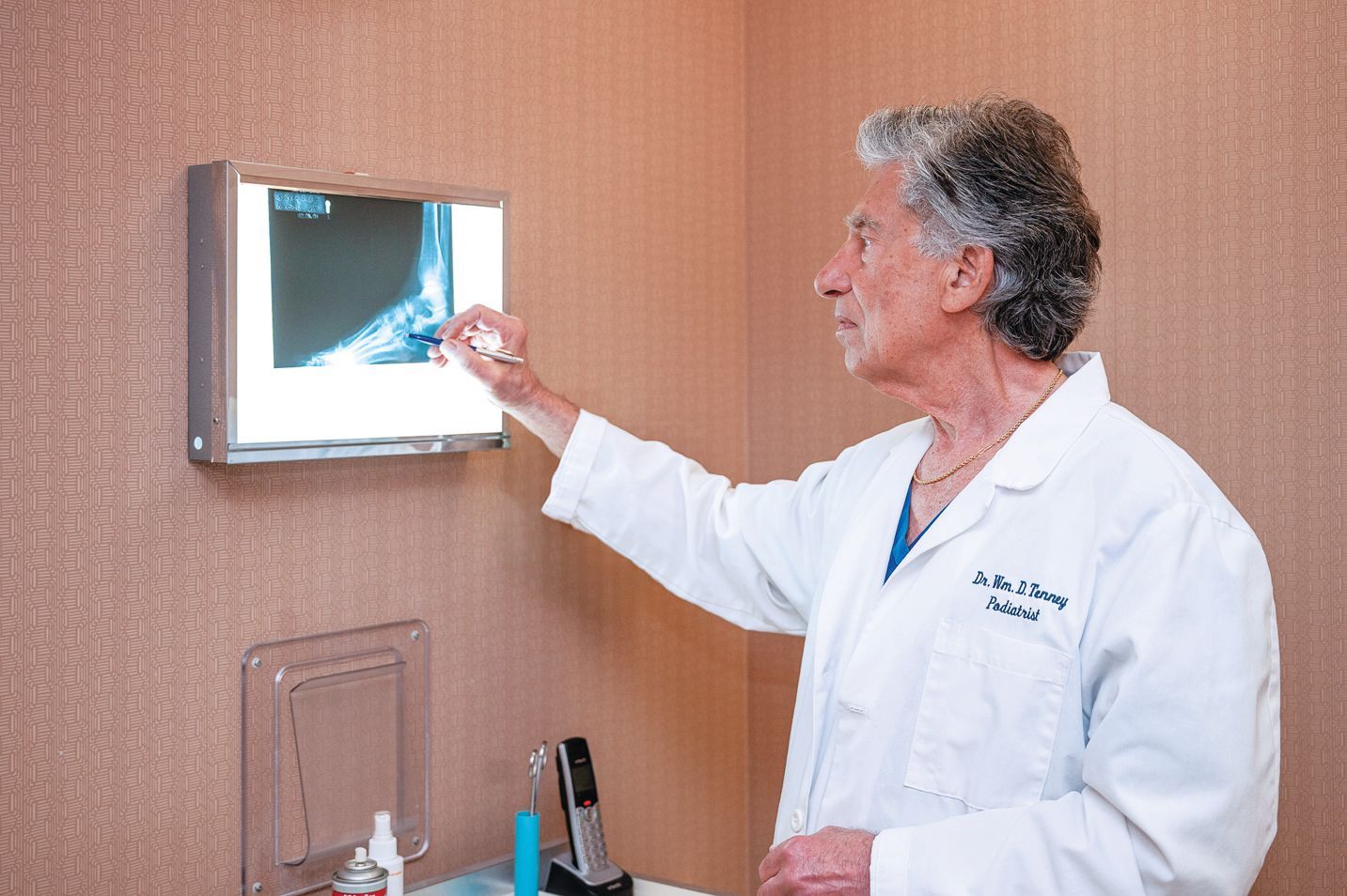 A physician in a white coat points to an X-ray of a foot on a wall-mounted light box in a clinical setting.