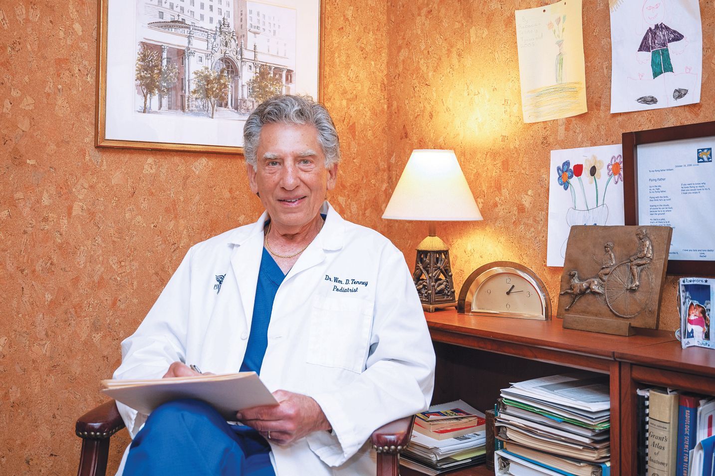 A medical professional in a white lab coat sitting in an office, holding a clipboard and looking toward the camera.