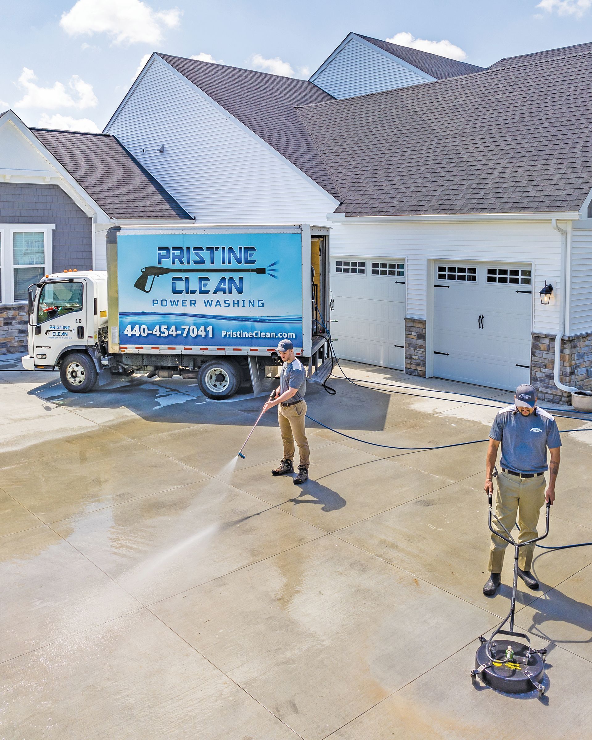 Two workers in uniform power wash a residential concrete driveway next to a Pristine Clean service truck.