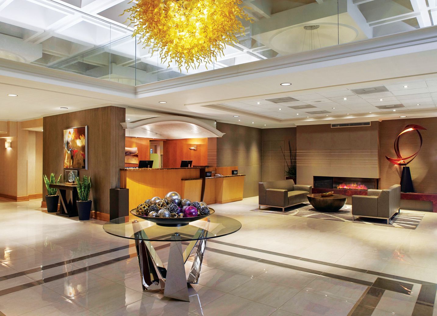 Modern hotel lobby with a reception desk, a central glass table with decor, comfortable seating, and a yellow chandelier.