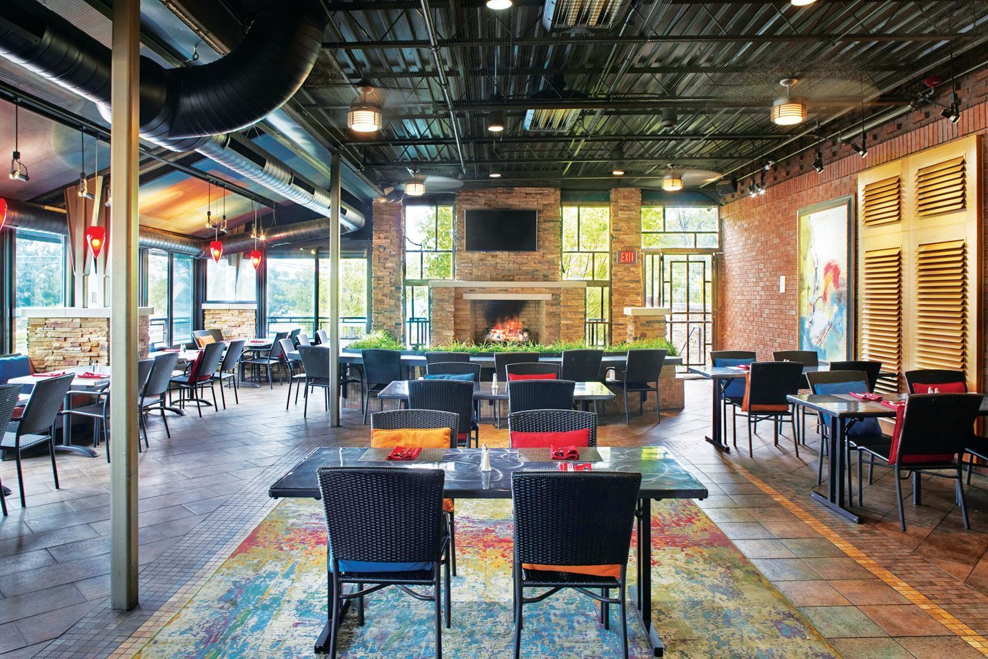 Indoor restaurant dining area with a brick wall, fireplace, patterned rug, and tables with black chairs.