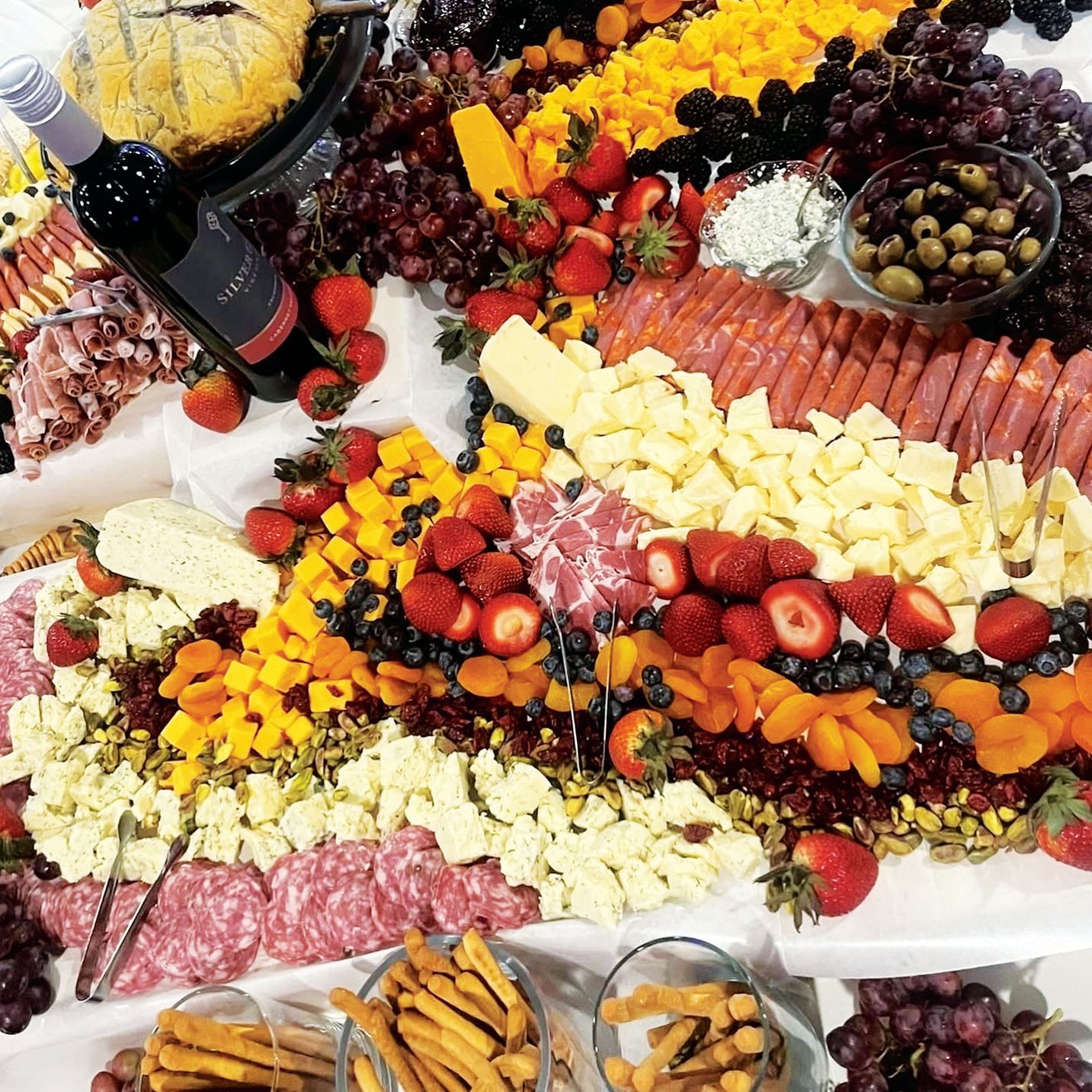 A large, abundant charcuterie board featuring various cheeses, meats, fruits, nuts, olives, and breadsticks.