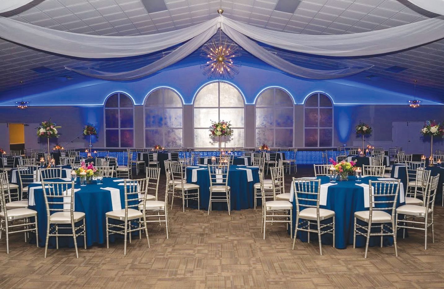 A banquet hall with tables covered in dark blue tablecloths, silver chairs, and white ceiling drapery with blue lighting.
