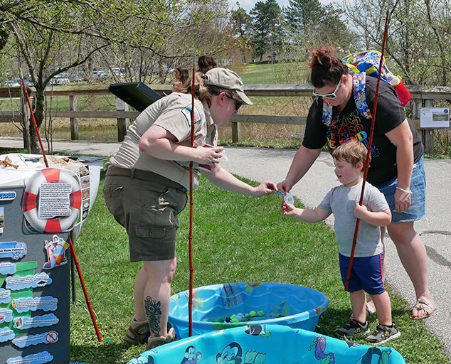 A park staffer assists a child using a makeshift fishing pole to reach into a blue plastic kiddie pool outdoors.