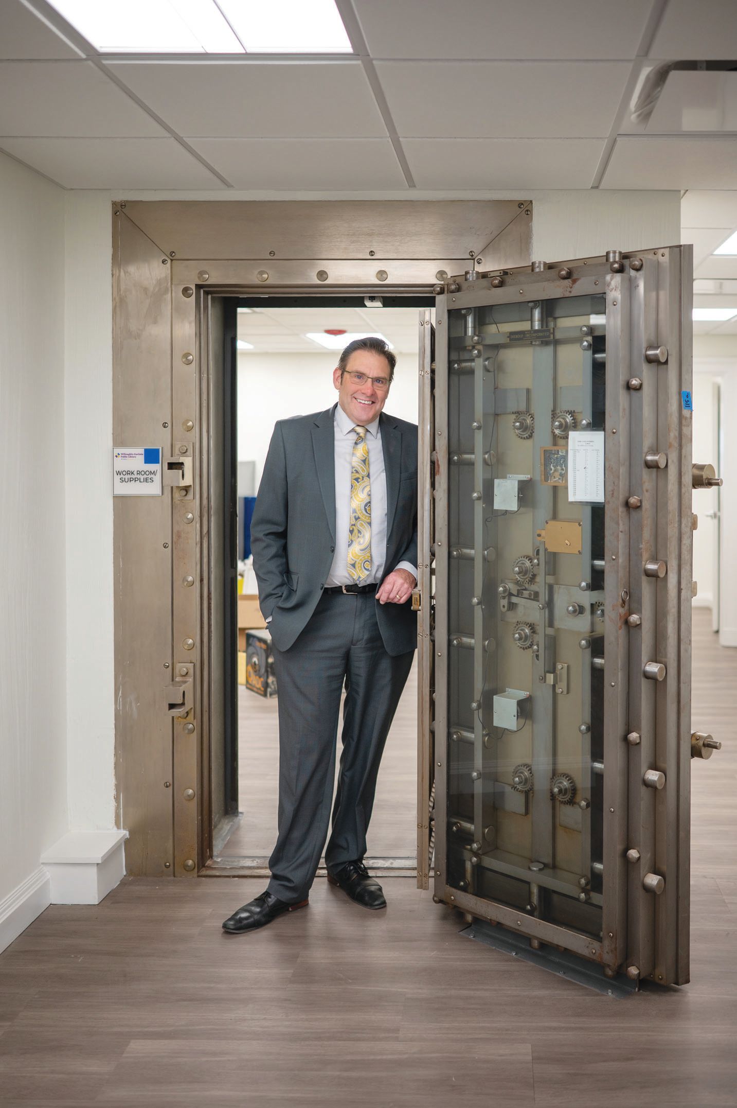 A professional in a grey suit and tie stands in the doorway of a large, open bank vault.