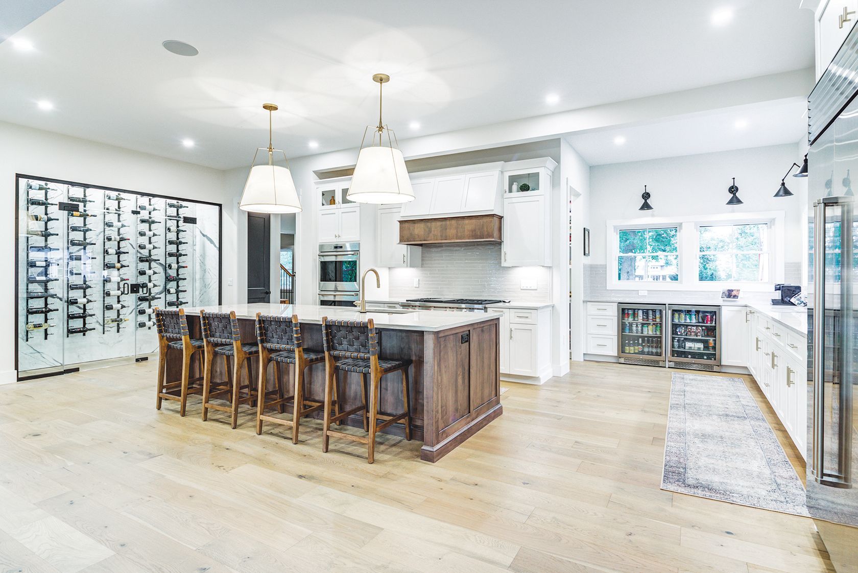 Modern kitchen with a wooden island, bar stools, white cabinetry, and a glass-enclosed wine rack on light wood floors.