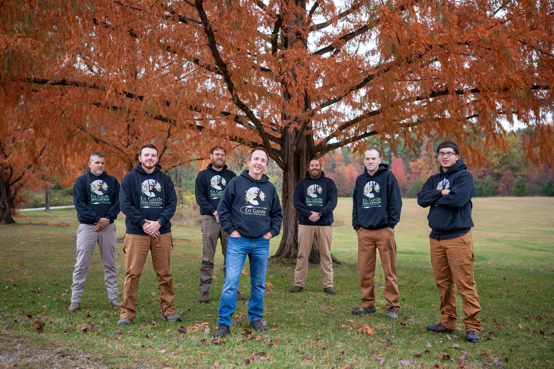 Seven people in matching dark hoodies and work pants pose in a line before a large, vibrant autumn tree.