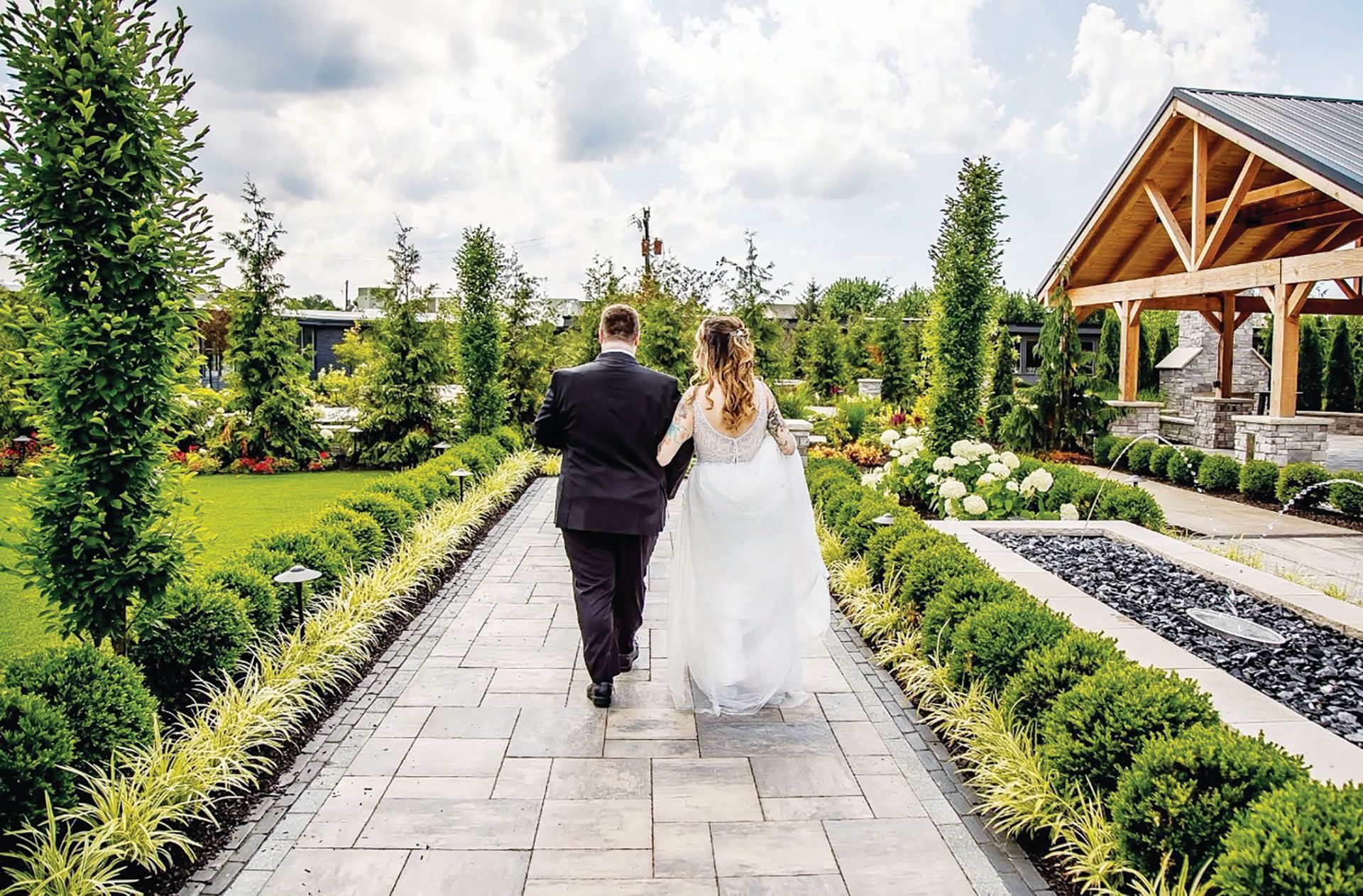 A bride and groom walk hand-in-hand down a stone path in a garden toward a wooden open-air structure.