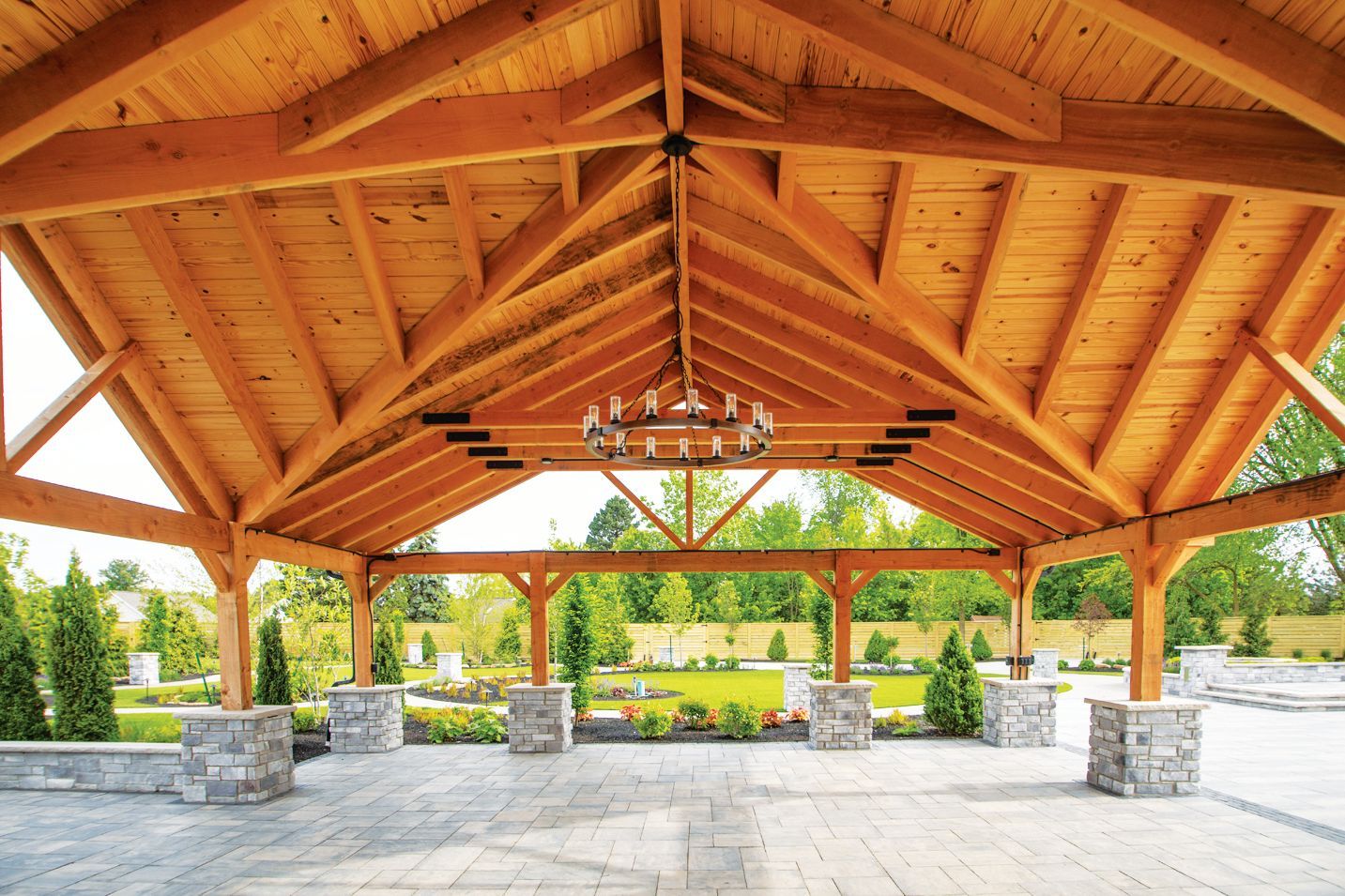 An open-air wooden pavilion with a vaulted ceiling, stone pillars, and a chandelier, overlooking a landscaped garden.