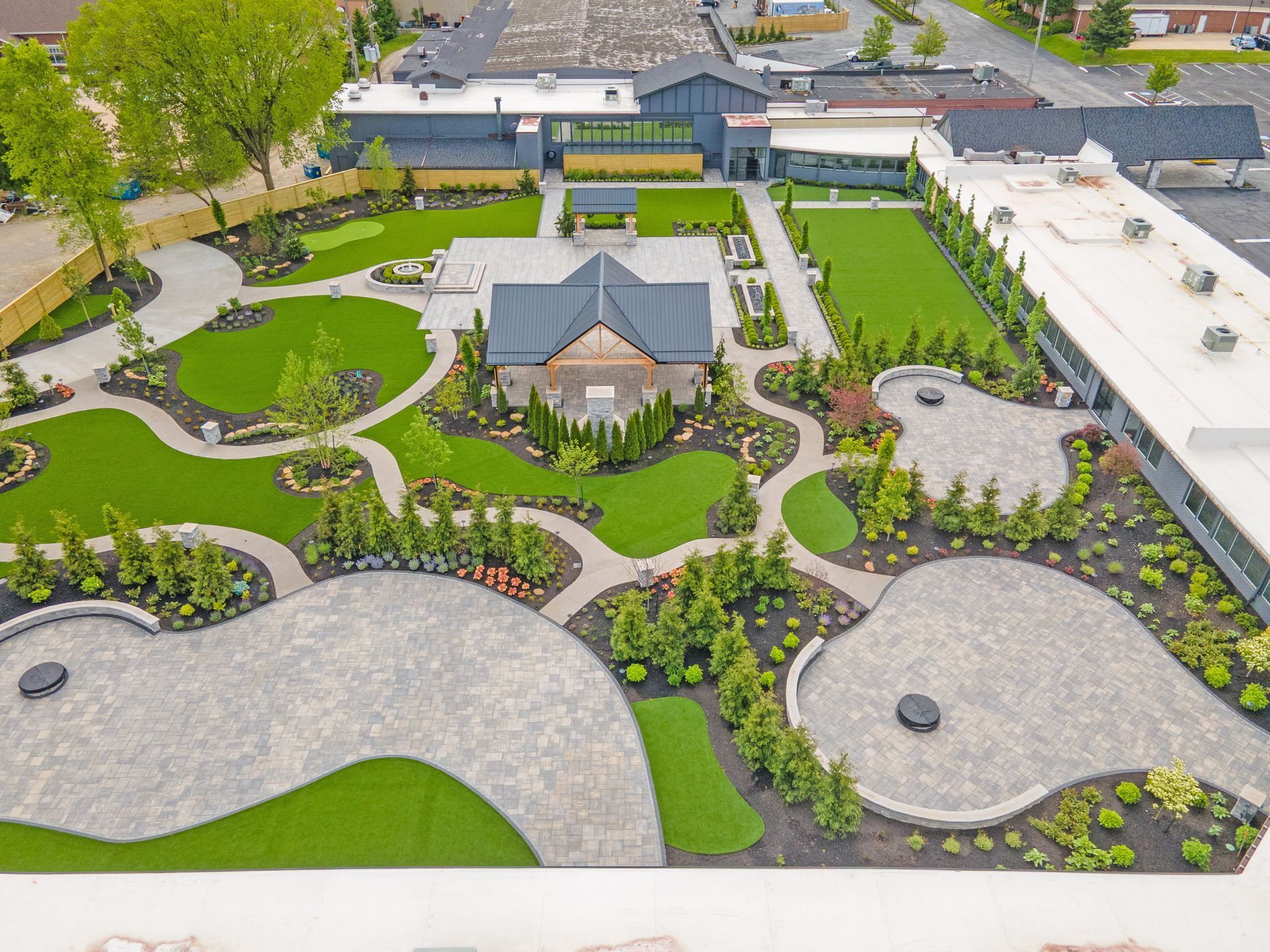 An aerial view of a landscaped park with winding stone paths, green lawns, decorative shrubs, and a central gazebo.
