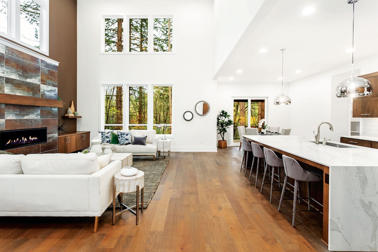 A modern, open-concept living room and kitchen with white walls, high ceilings, wood floors, and neutral furniture.
