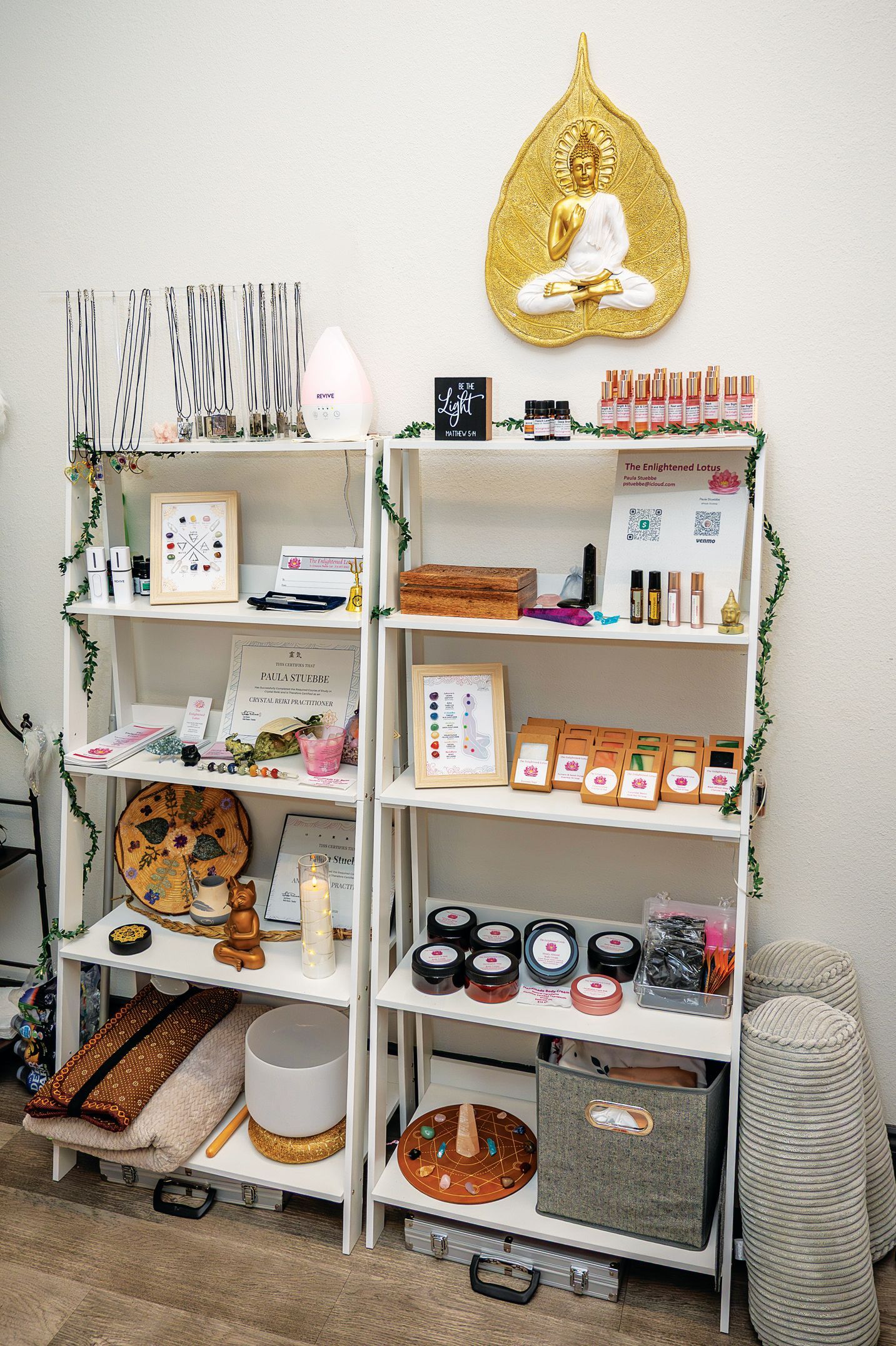 Two white shelving units filled with various wellness products, crystals, and decor, topped by a gold Buddha wall plaque.