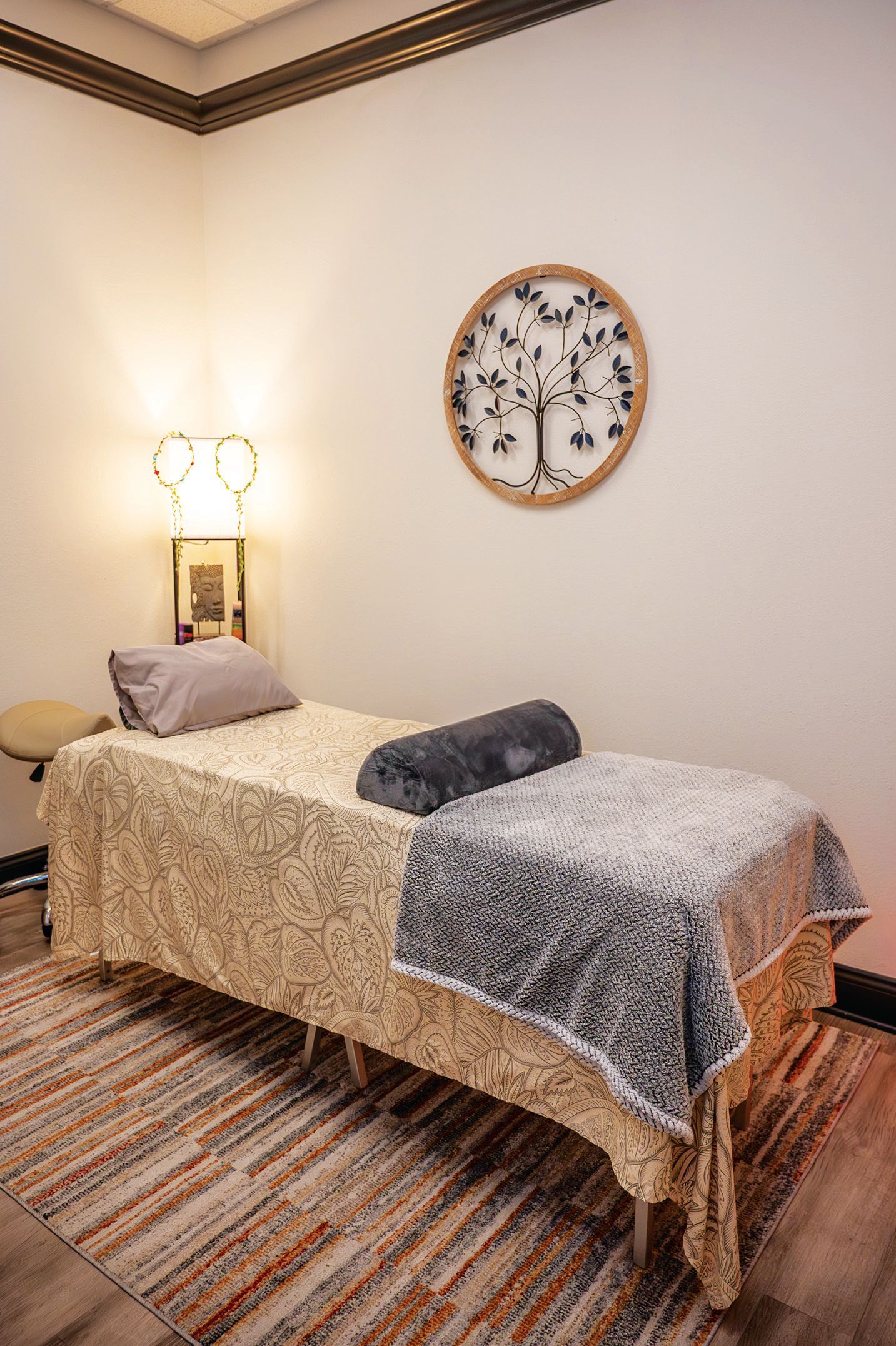 A massage table with a textured cream cover, a grey blanket, and a pillow, positioned in a quiet room with wall art.
