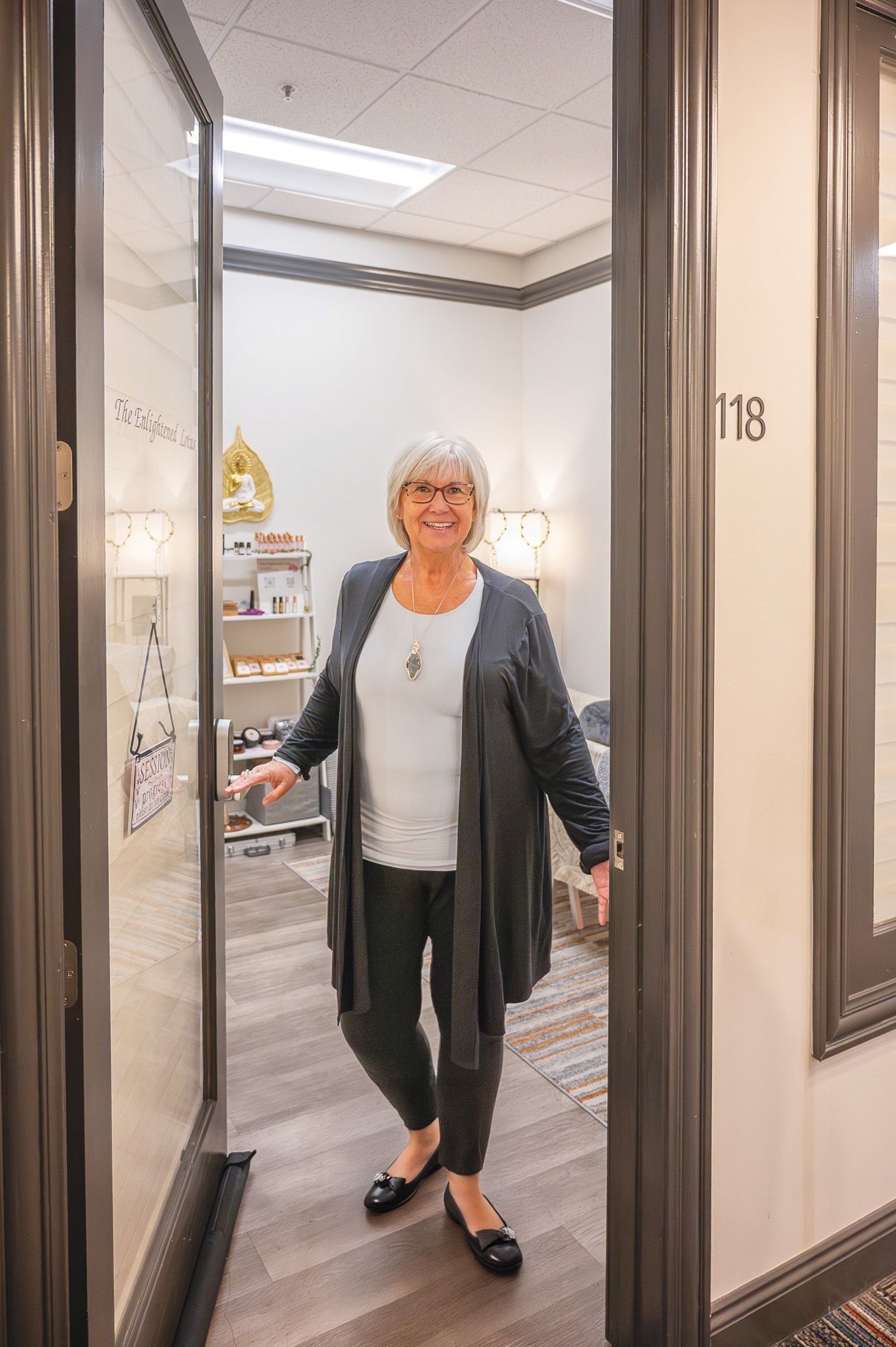 A smiling person wearing a light top and dark cardigan stands in the doorway of office 118, holding the door open.