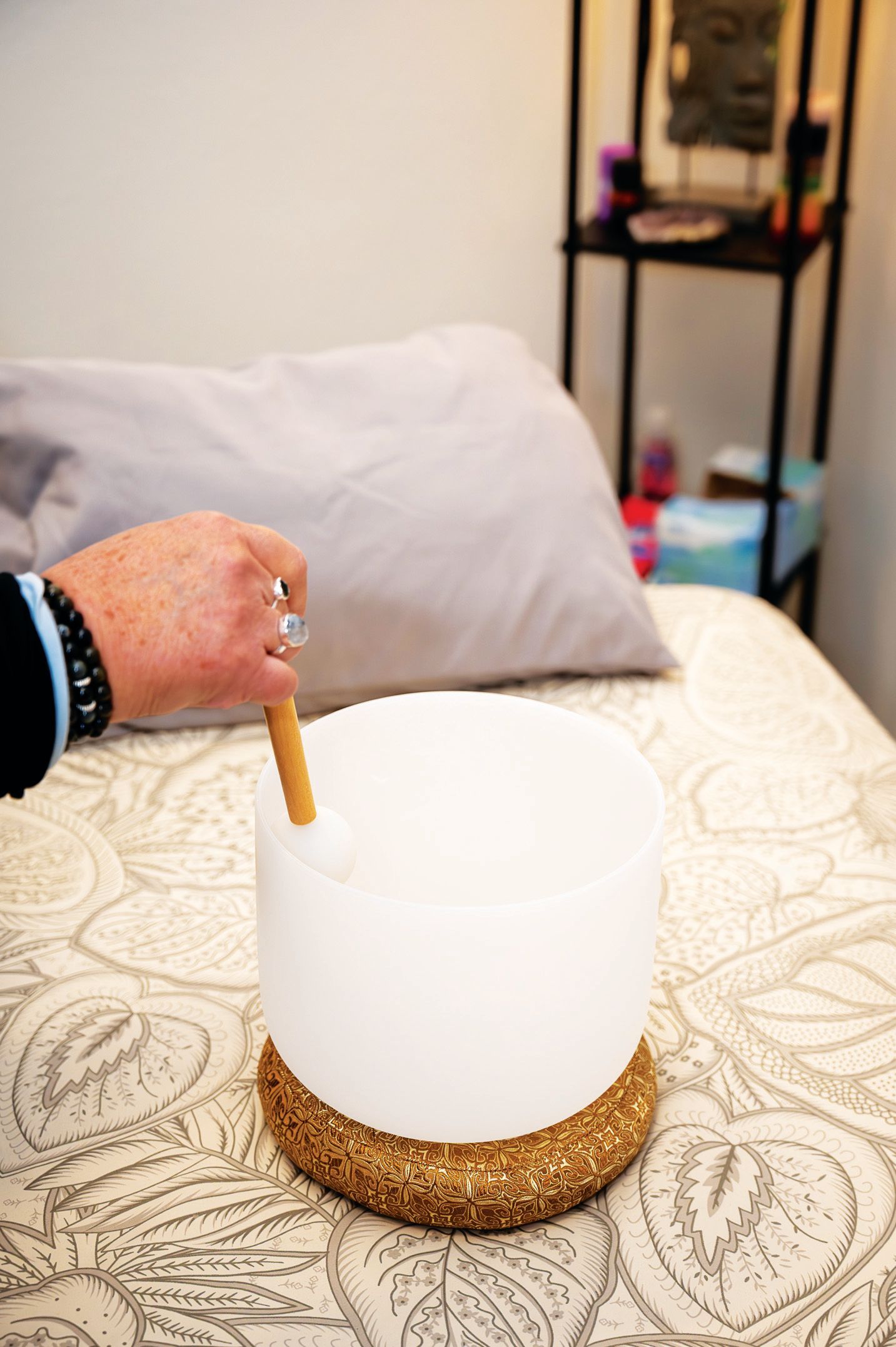 A hand uses a mallet to strike a white, translucent crystal singing bowl resting on a textured base on a patterned bed.