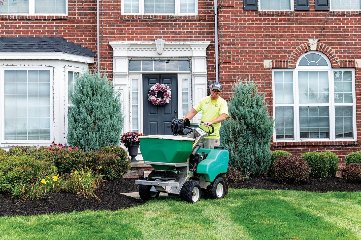 A person in a neon yellow shirt operates a green fertilizer spreader on a front lawn in front of a brick house.