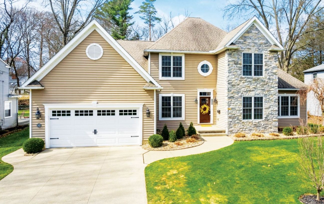 A two-story suburban home with tan vinyl siding, a stone facade, a two-car garage, and a lush green lawn.