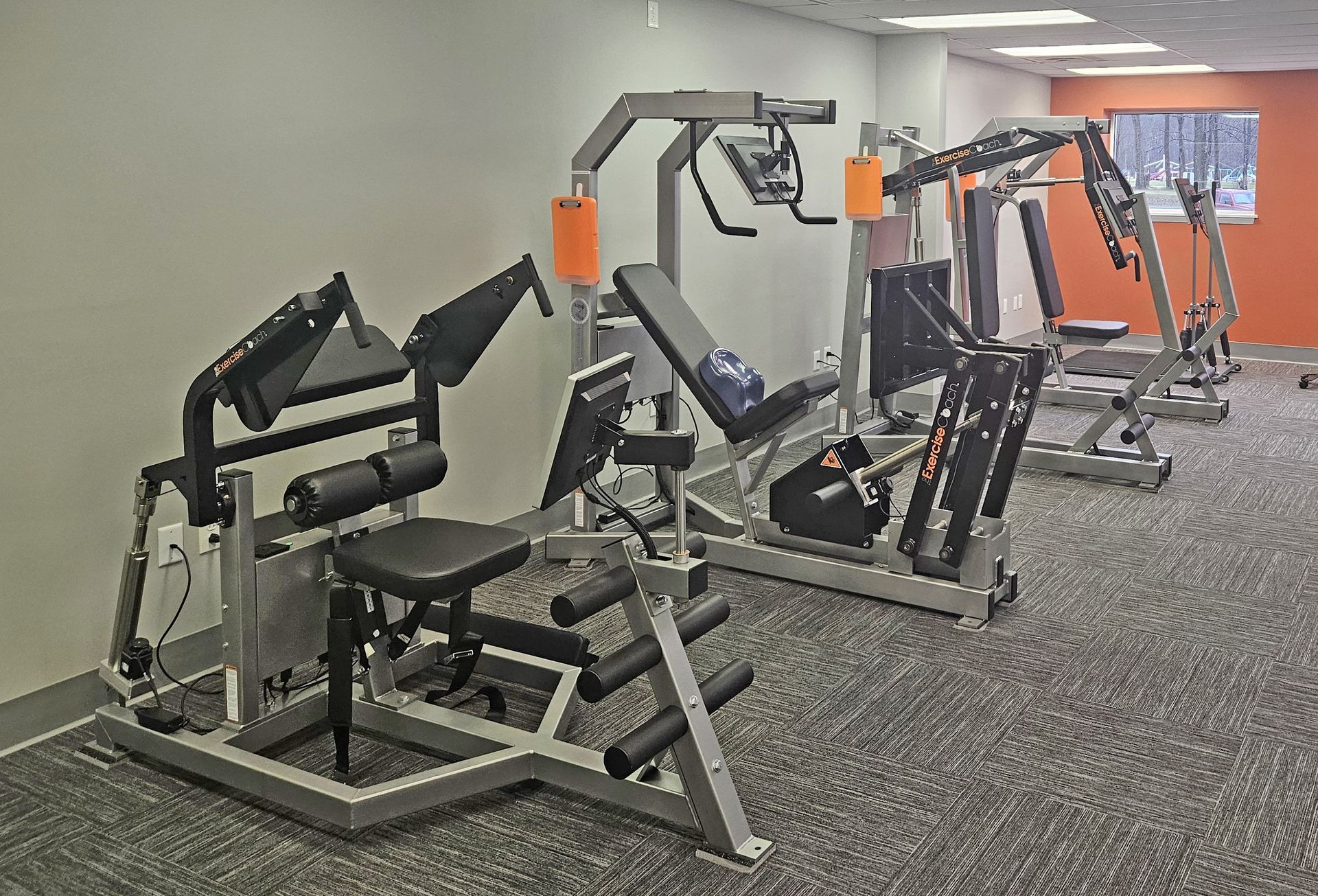 A gym room with multiple stationary weight training machines arranged in a row on a gray carpeted floor.