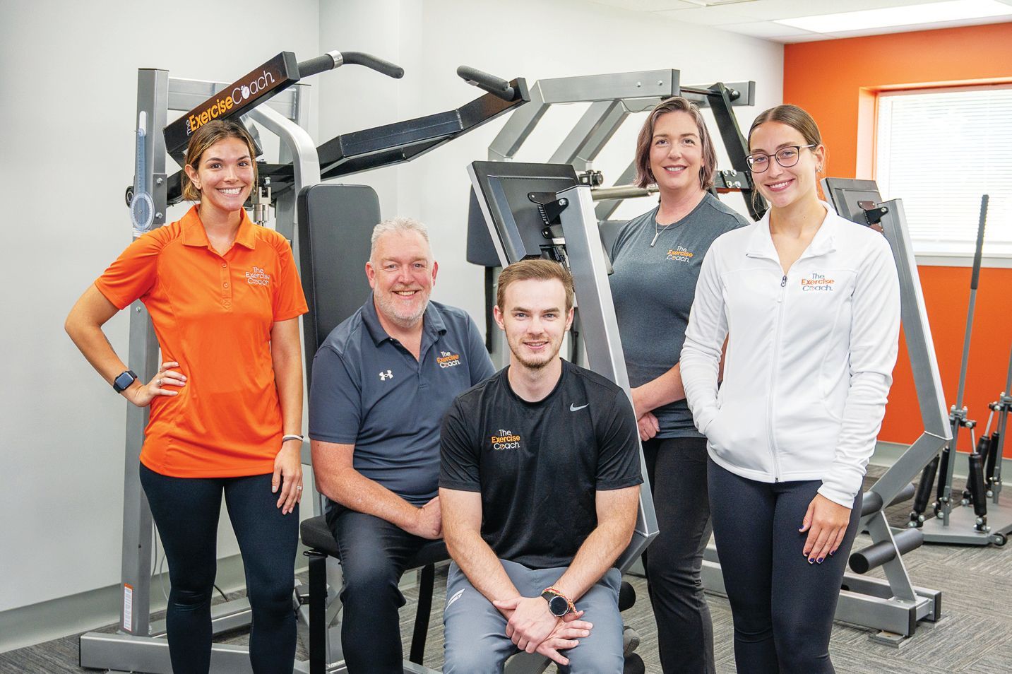 Five people standing and sitting in a gym setting with workout equipment in the background.