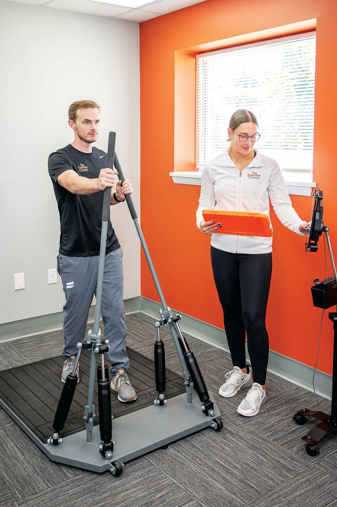 A person stands on a balance testing platform while holding handles, and an observer holds a tablet to record data.