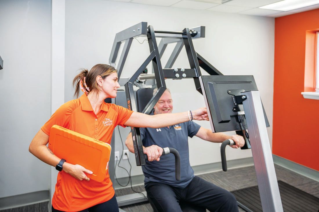 A fitness professional in an orange shirt guides a person using a piece of strength-training equipment in a gym.