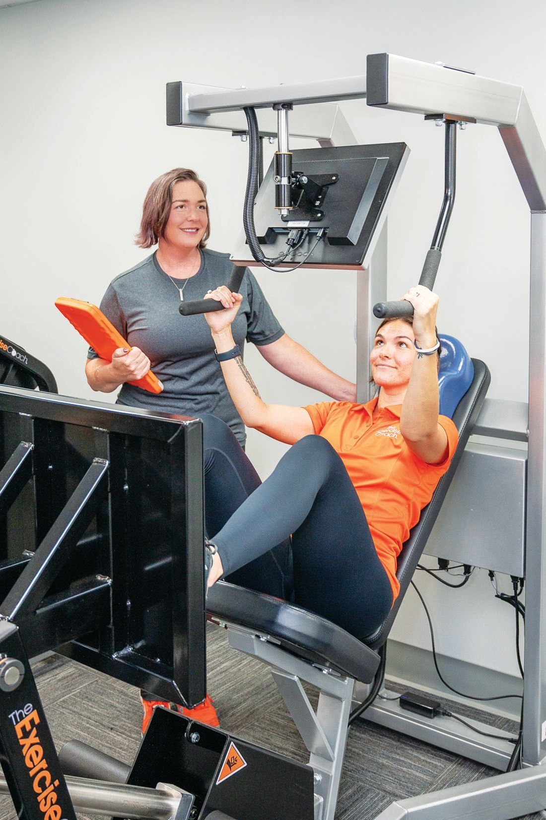 A professional trainer supervises an individual using a seated leg press machine in a fitness facility.