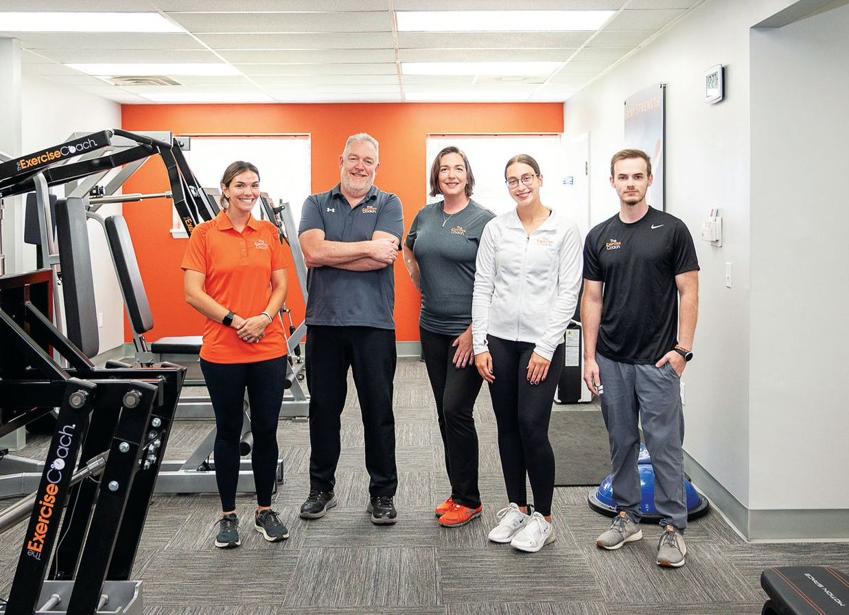 Five people stand in a fitness facility with gym equipment, smiling towards the camera.