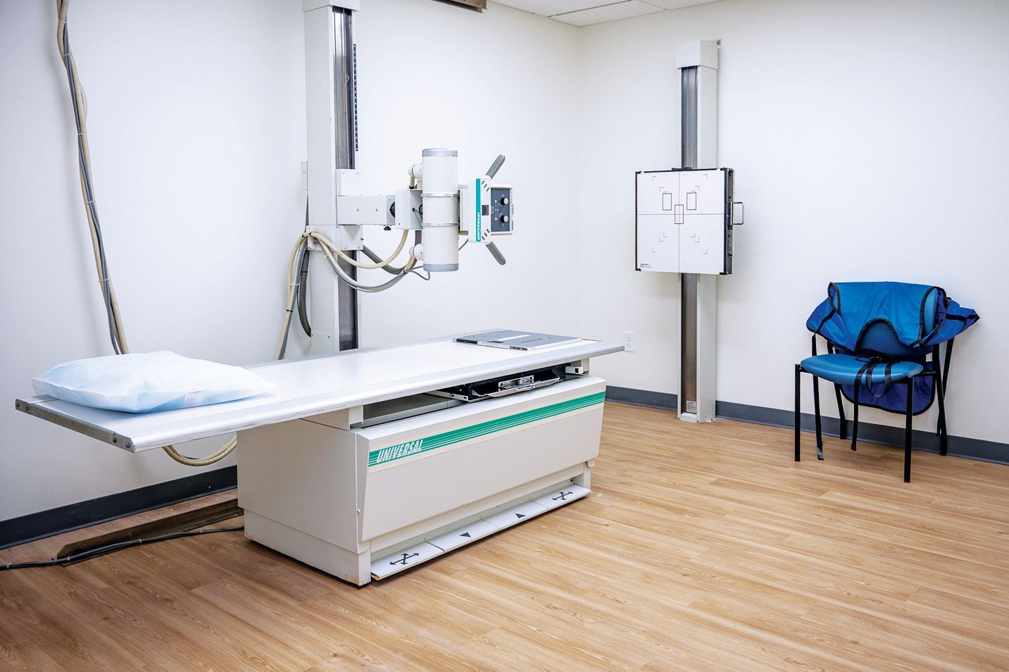 A medical imaging room with an X-ray machine, a patient table, and a blue chair on a wood-look floor.