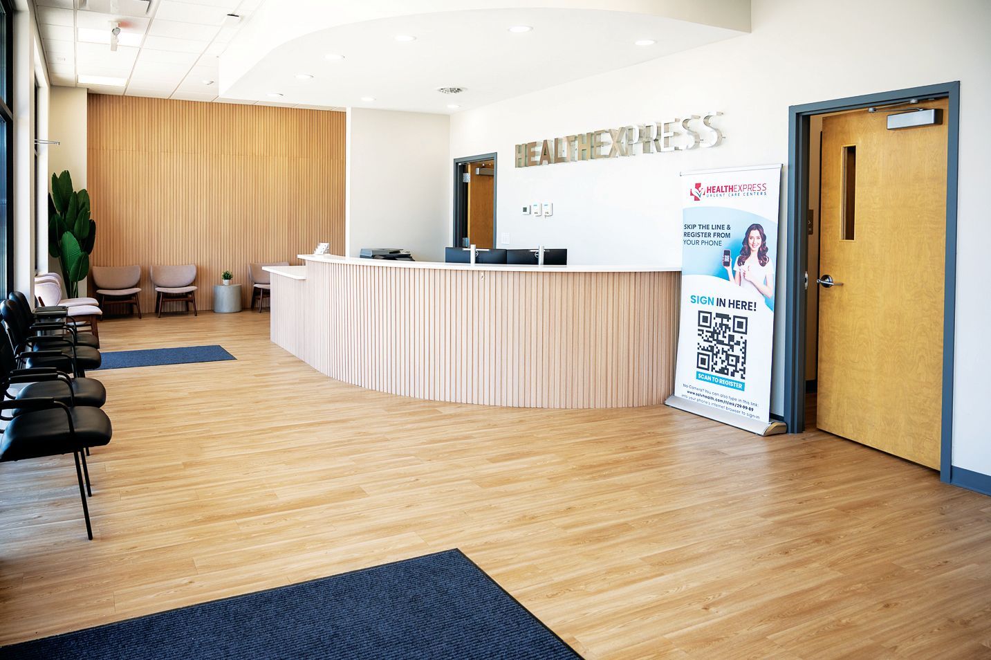 Reception area with a curved wooden desk, light wood walls, black chairs, and a vertical sign with a QR code by a doorway.