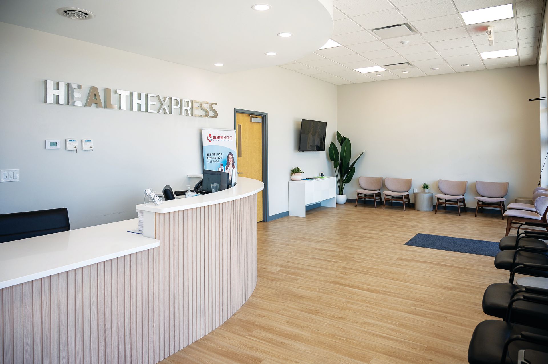 A modern medical reception area with a curved desk, signage, waiting chairs, and wood-look flooring.