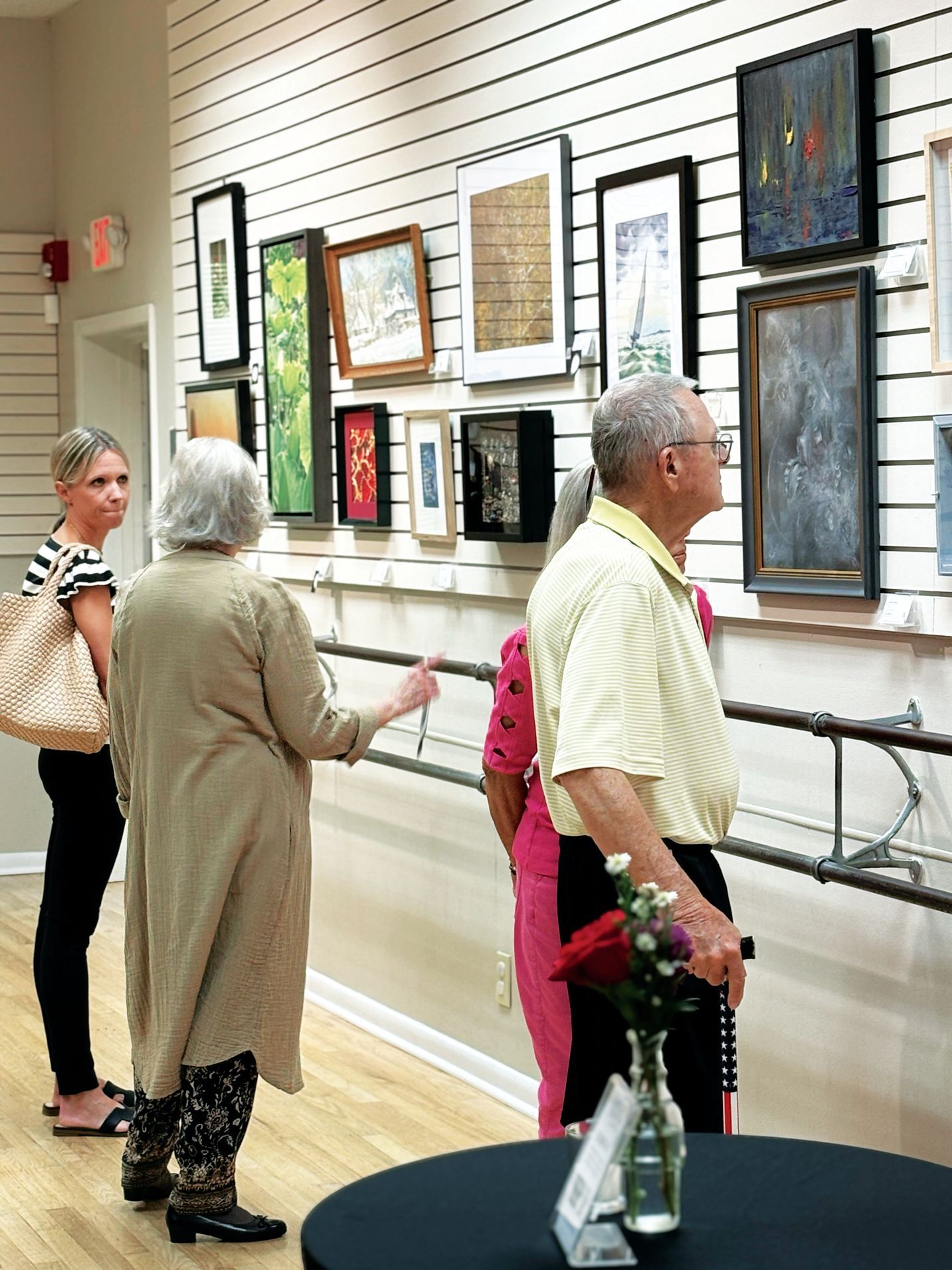 A group of people observe an art gallery display featuring framed paintings mounted on a slatted wall.