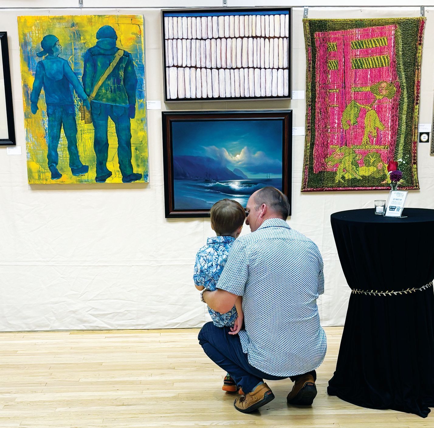 A person holding a child while crouching to look at a dark, atmospheric painting in a bright art gallery.
