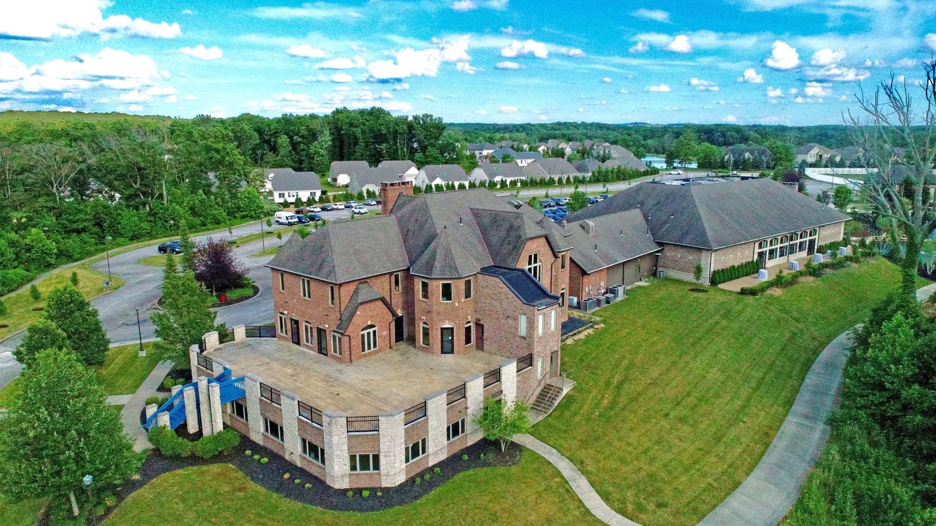 Aerial view of a large, multi-story red brick building with a gray shingled roof, surrounded by green lawns and trees.