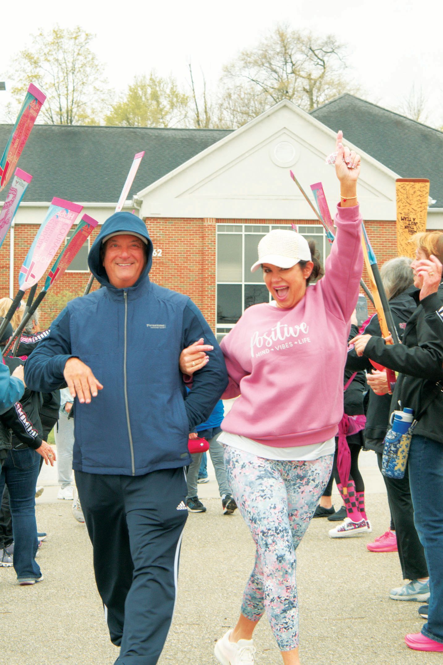 A man in a blue hoodie and a woman in a pink sweatshirt and patterned leggings walk arm-in-arm, smiling, outdoors.