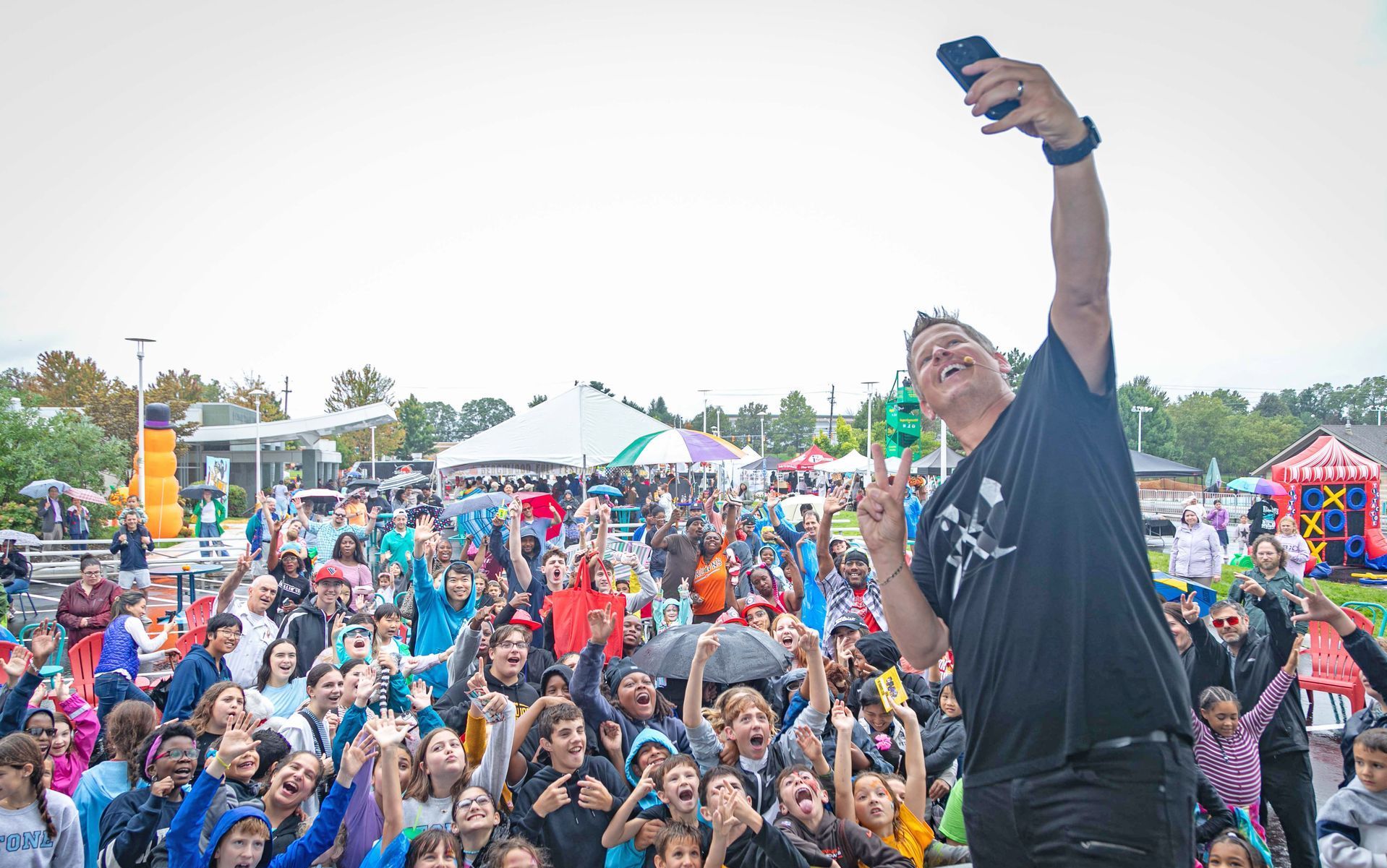 A person takes a selfie with a large, diverse crowd outdoors at a festival, with tents and decorations in the background.