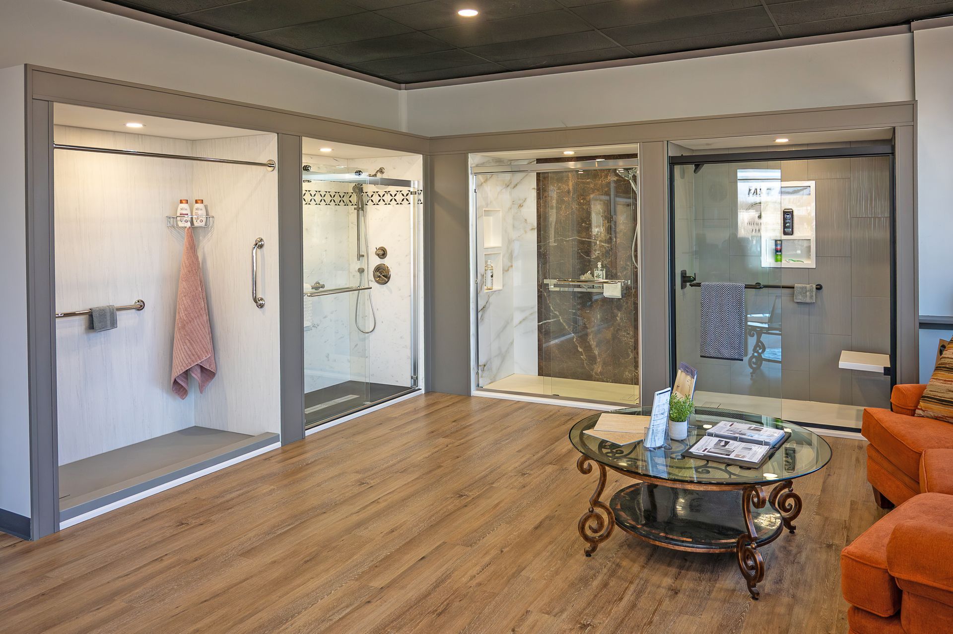 A showroom features three walk-in shower displays alongside a glass coffee table and orange seating on a wood-look floor.