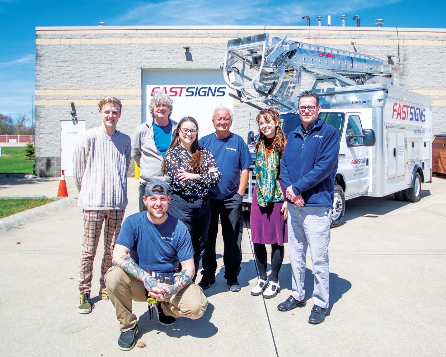 A team of seven people pose for a photo in front of a white FastSigns service truck parked at an industrial building.