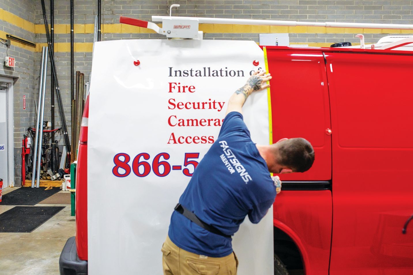 A person in a blue t-shirt applies a white vinyl decal with red text to the side of a red utility van in a workshop.