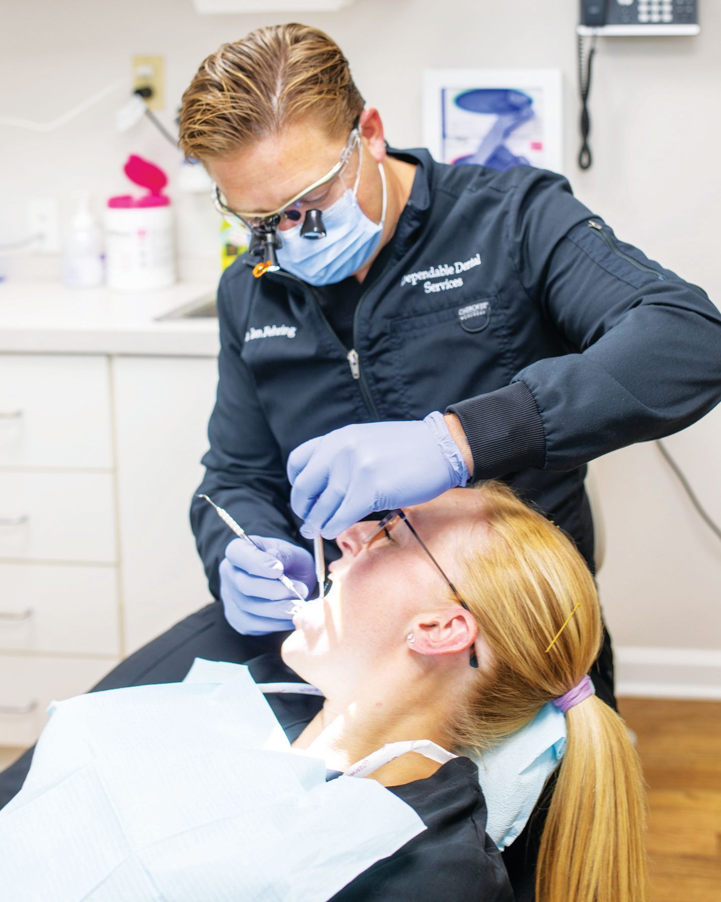 A dentist wearing a mask and magnifying glasses examines a patient's teeth in a clinical setting.