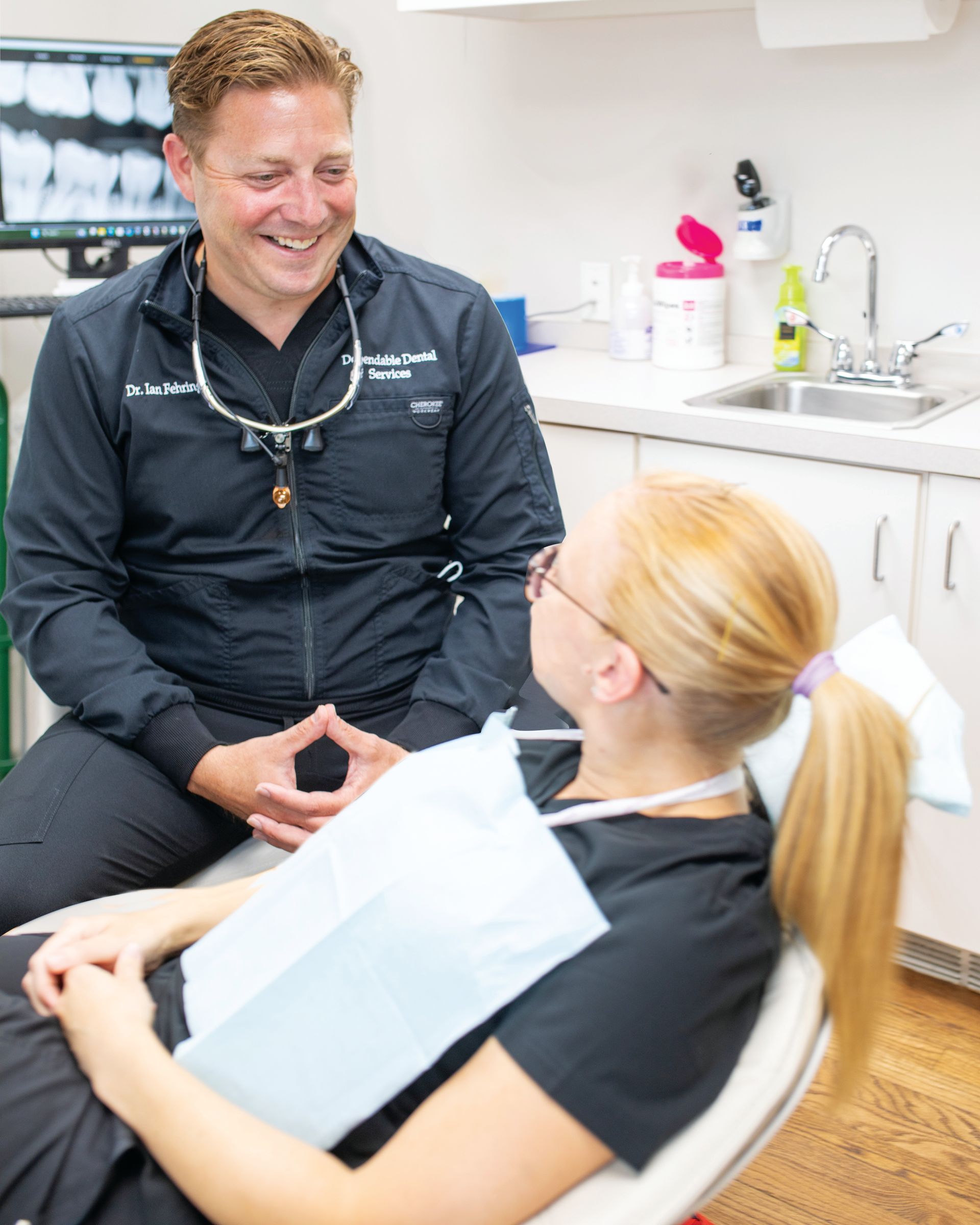 A smiling dentist in scrubs and magnifying loupes talks to a patient sitting in a dental chair in an office.