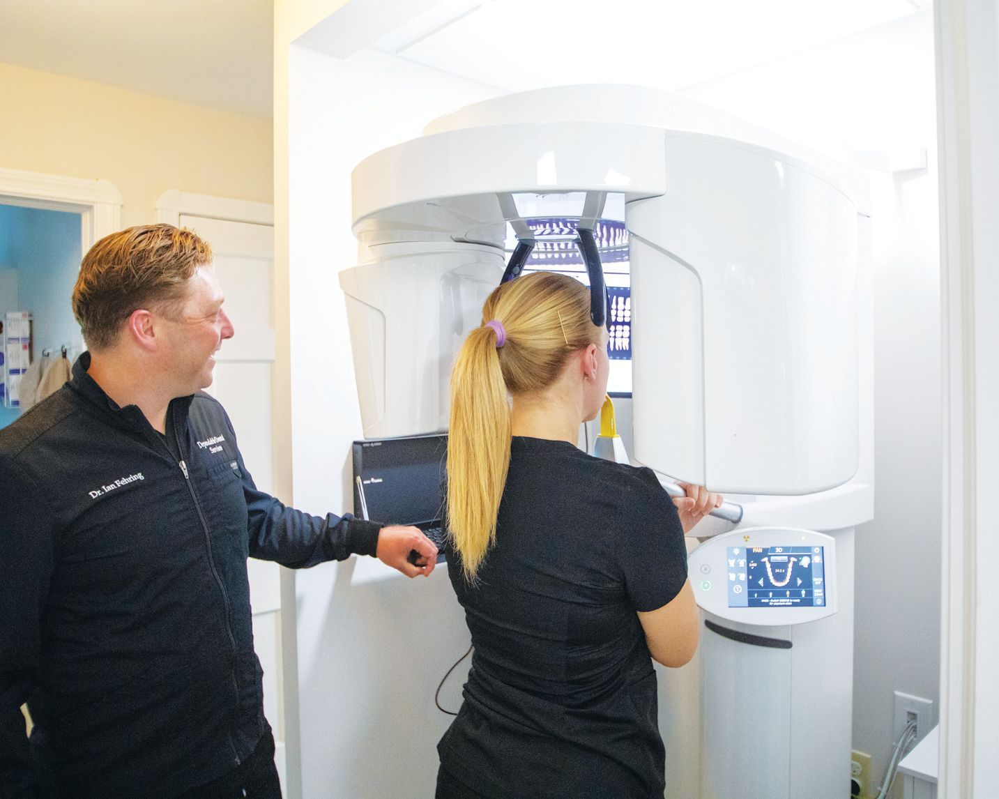 A technician assists a person with a dental X-ray machine in a clinical office setting.