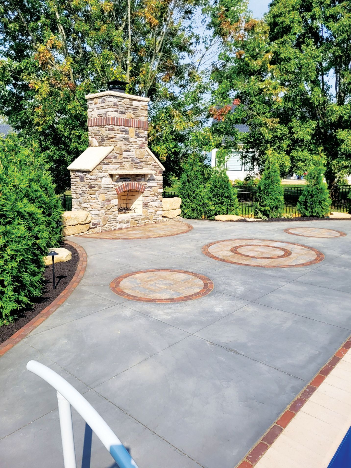 An outdoor stone fireplace sits on a gray patio featuring three circular inlay patterns, surrounded by lush green trees.